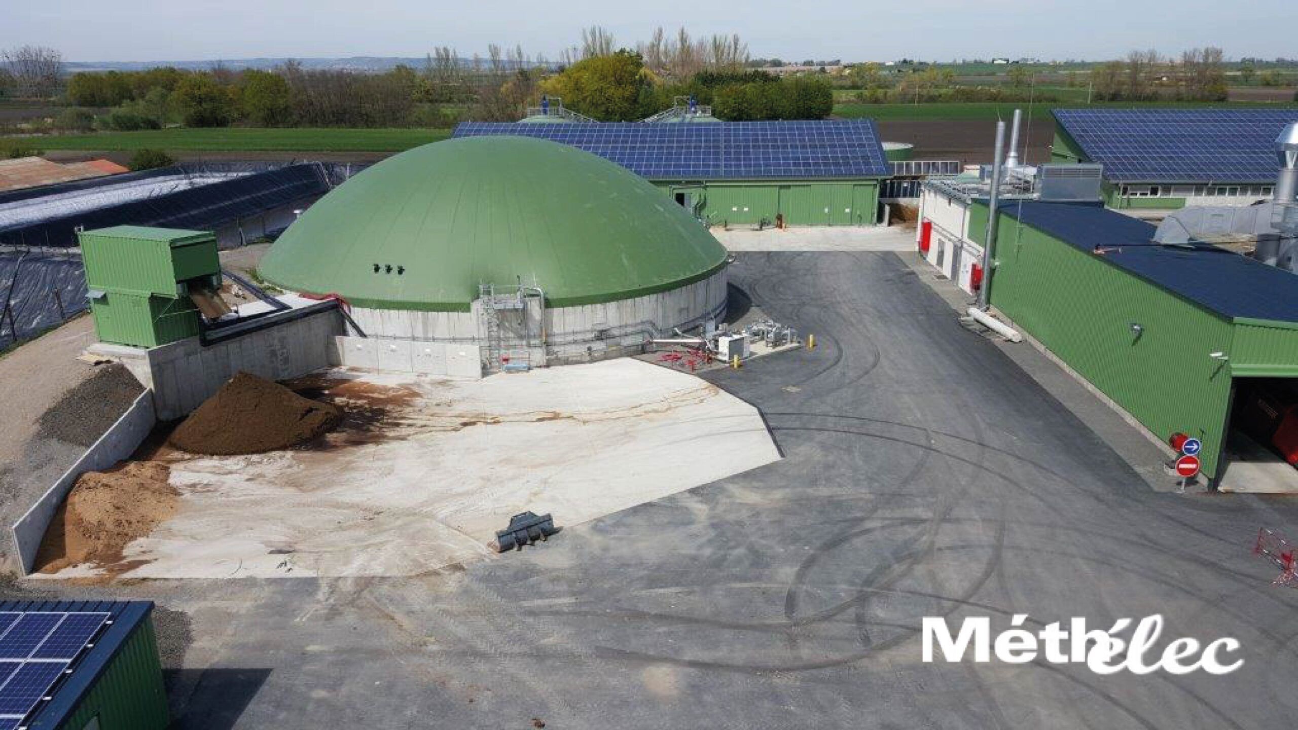 Vue aérienne d’un site de méthanisation Méthélec avec digesteur vert, bâtiments industriels et panneaux photovoltaïques.